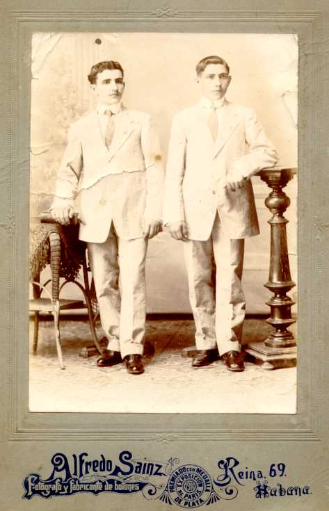 Foto en la que se ve a dos jovenes indianos (16-18 años) vestidos de traje blanco con corbata clara, tiene marco de cartulina que dice Alfredo Sainz fotografo y fabricante de botones, Reina 69 Habana